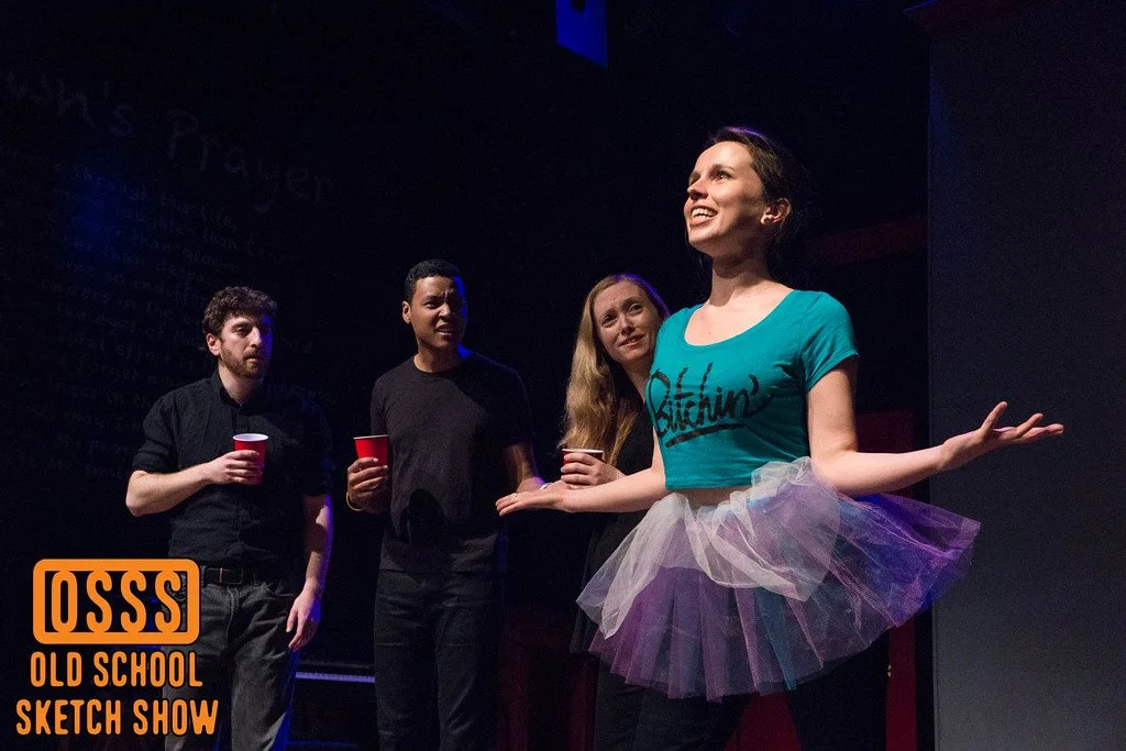 Kate Shine, a young white woman wearing a turquoise t-shirt and a tutu, performs in Old School Sketch Show, with three other actors dressed in black.