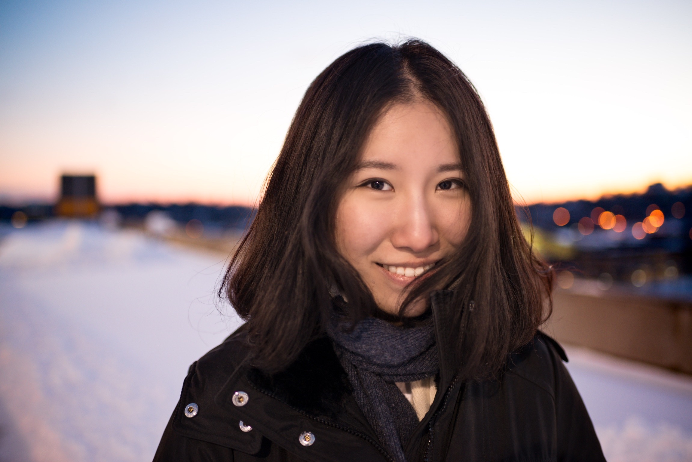Image of a young Asian woman with long shoulder length hair standing in front of a body of water, out of focus and at sunset. She is wearing a dark jacket and scarf and staring into the camera, smiling.