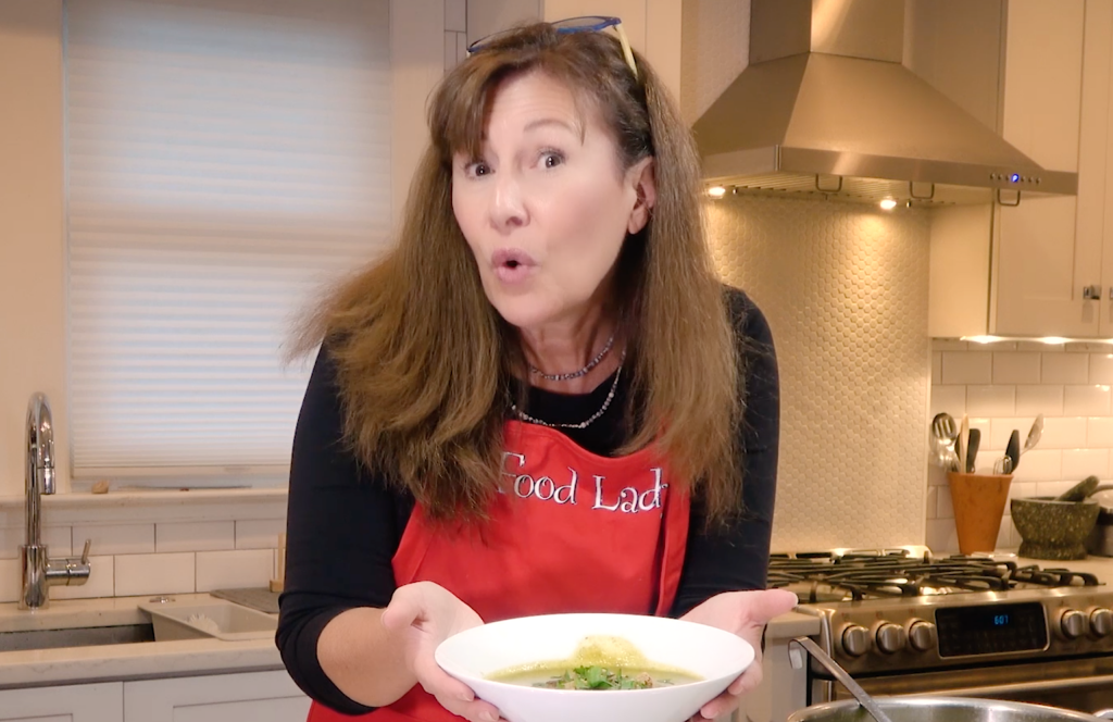 A woman in a red "Food Lady" apron holds a bowl of soup in a modern kitchen, displaying excitement.
